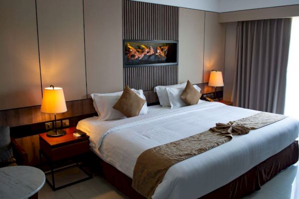 A cozy hotel room featuring a large bed with brown accents, bedside tables with lamps, and a wall-mounted fireplace.