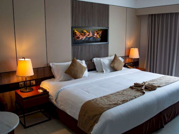 A cozy hotel room featuring a large bed with brown accents, bedside tables with lamps, and a wall-mounted fireplace.