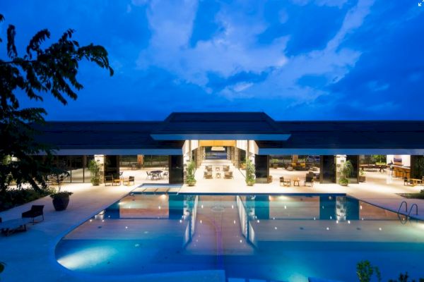 A luxurious pool area at night, featuring an expansive building with open spaces and well-lit seating arrangements under a twilight sky.