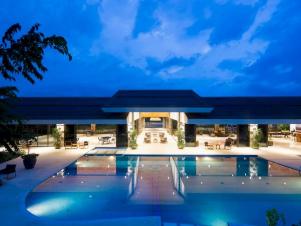 A luxurious pool area at night, featuring an expansive building with open spaces and well-lit seating arrangements under a twilight sky.