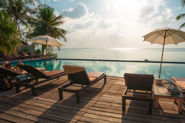 A serene beach scene featuring lounge chairs, umbrellas, palm trees, and an infinity pool overlooking a calm ocean under a partly cloudy sky.
