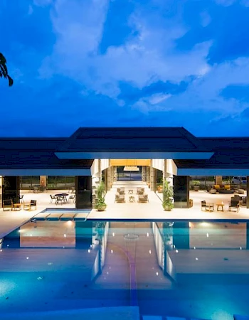 This image depicts a luxurious outdoor pool and a modern building with open seating areas, lit beautifully in the evening.