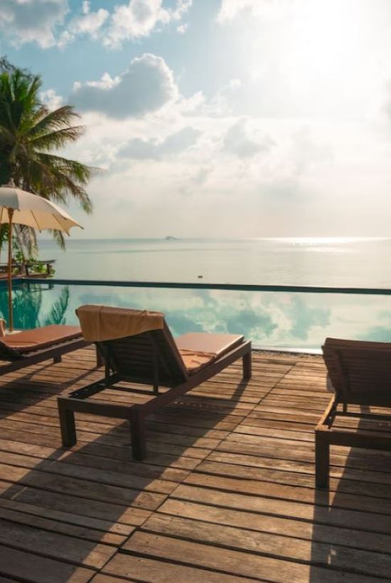A serene poolside setting with wooden lounge chairs, umbrellas, and palm trees, overlooking a calm body of water under a partly cloudy sky.