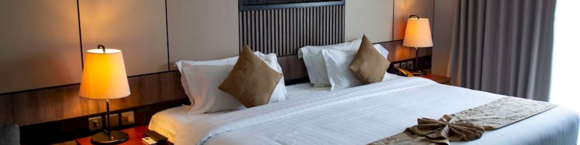 A cozy hotel bedroom with a king-sized bed, bedside tables with lamps, wall art, and a window with curtains; warm and inviting ambiance.