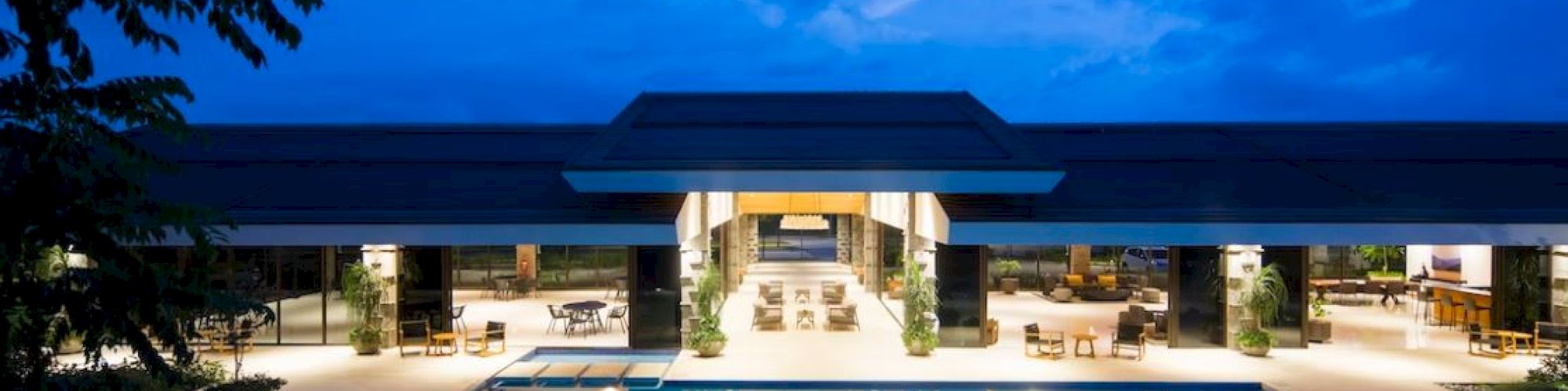A luxurious modern building with an illuminated swimming pool and outdoor seating area at dusk, surrounded by trees and a blue sky.