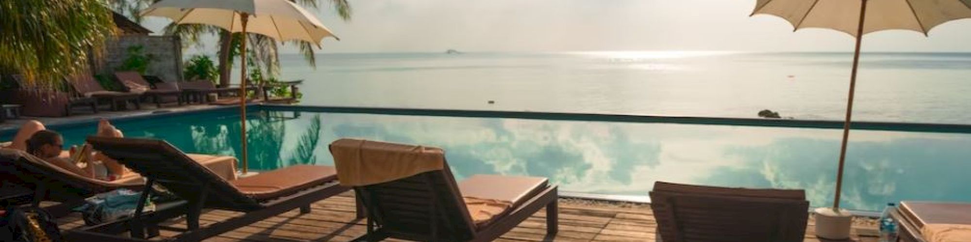 The image shows a serene poolside scene with lounge chairs, umbrellas, palm trees, and a beautiful horizon over a calm body of water.