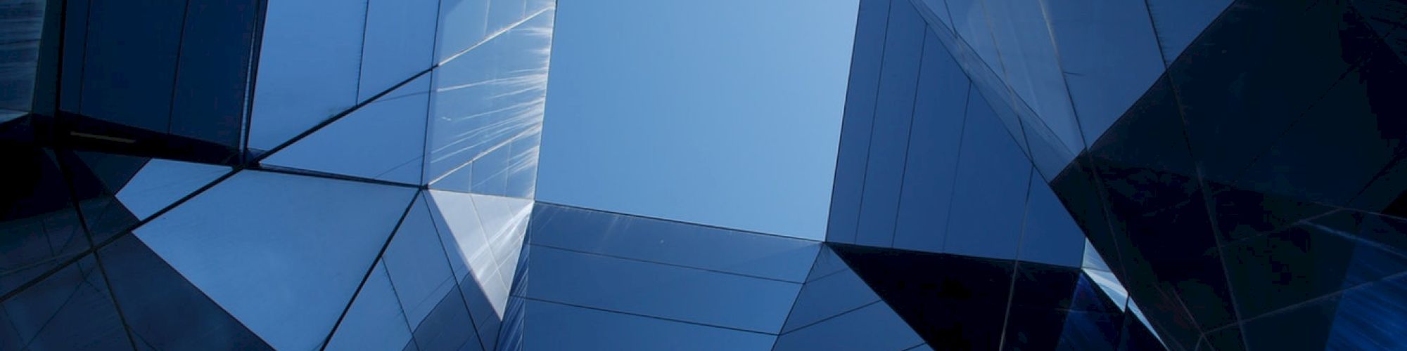 The image depicts a view looking up at a geometric arrangement of reflective glass panels, with a clear blue sky visible in the center of the composition.