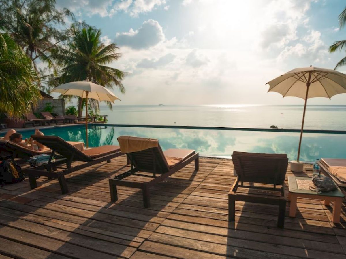 A serene poolside scene with lounge chairs, beach umbrellas, and palm trees under a bright sky, overlooking a calm ocean.