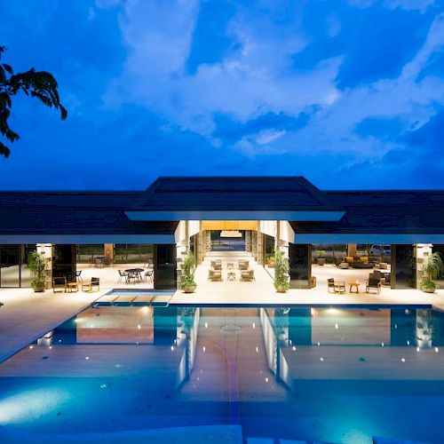 The image shows a modern, brightly lit building with a large, serene swimming pool in the foreground, creating a luxurious ambiance.