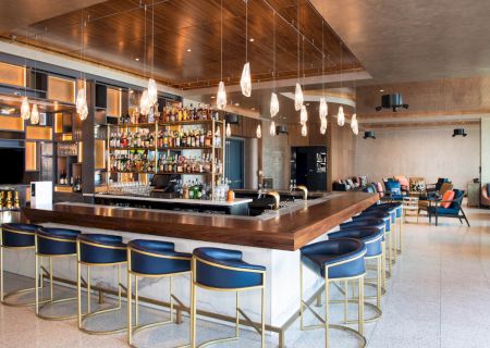 A modern bar with a wooden counter, blue chairs, hanging lights, and numerous bottles on display. The seating area is visible in the background.