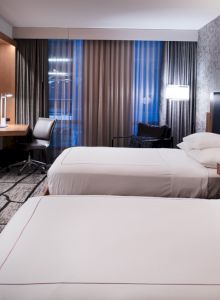 A modern hotel room with two beds, a wall-mounted TV, a desk with a chair, and stylish lighting, featuring a contemporary design.