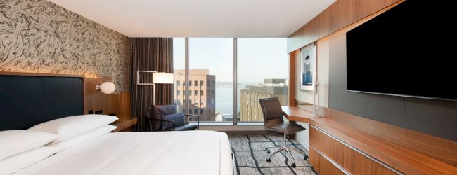 A modern hotel room featuring a large bed, wall-mounted TV, desk with chair, and floor-to-ceiling windows offering a view of city buildings.
