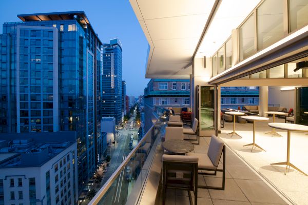 This image shows a modern cityscape with tall buildings and a spacious balcony at dusk, featuring tables, chairs, and city lights.