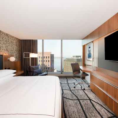 A modern hotel room with a large bed, a wall-mounted TV, a desk with a chair, and a view of buildings through a large window at the end.