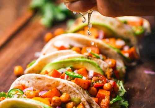 The image shows a hand squeezing a lime over a board of vegetable tacos, garnished with chickpeas, lettuce, and other fresh ingredients.