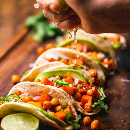 The image shows a hand squeezing a lime over a board of vegetable tacos, garnished with chickpeas, lettuce, and other fresh ingredients.