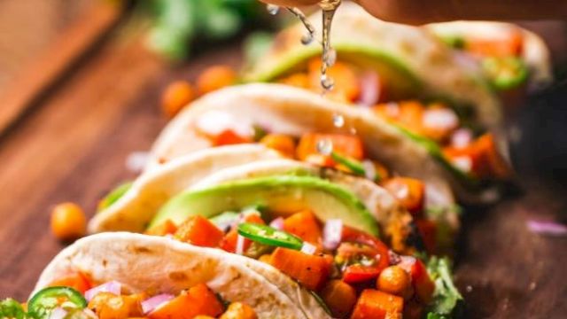 The image shows a hand squeezing a lime over a board of vegetable tacos, garnished with chickpeas, lettuce, and other fresh ingredients.