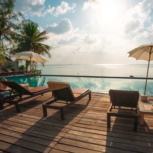 Lounge chairs and umbrellas are set around a serene infinity pool overlooking the ocean, with palm trees and a bright sky in the background.