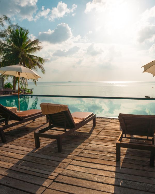 Lounge chairs and umbrellas are set around a serene infinity pool overlooking the ocean, with palm trees and a bright sky in the background.