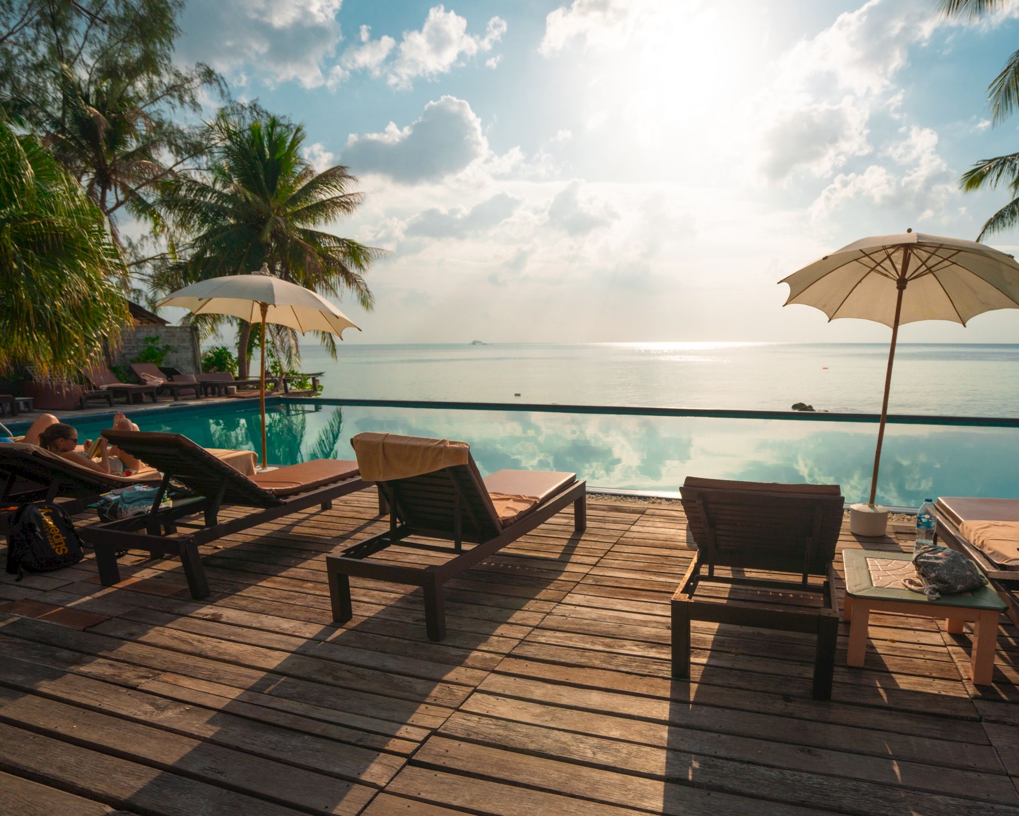 Sun loungers under umbrellas by an infinity pool, overlooking the ocean, with palm trees and a serene sky in the background.