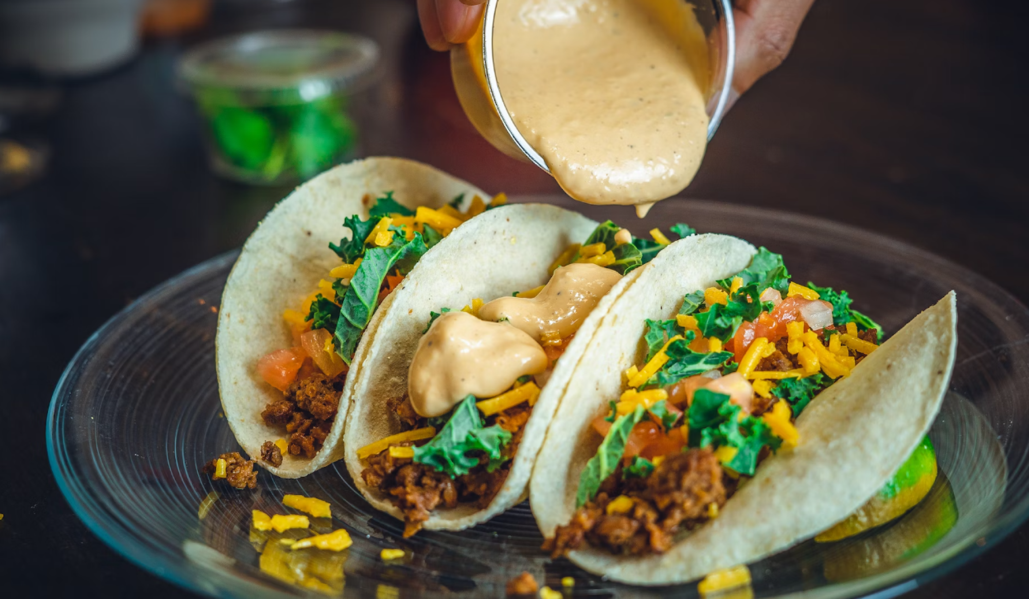 The image shows three tacos filled with meat, shredded cheese, tomatoes, and greens on a plate. A hand drizzles creamy sauce on one taco.