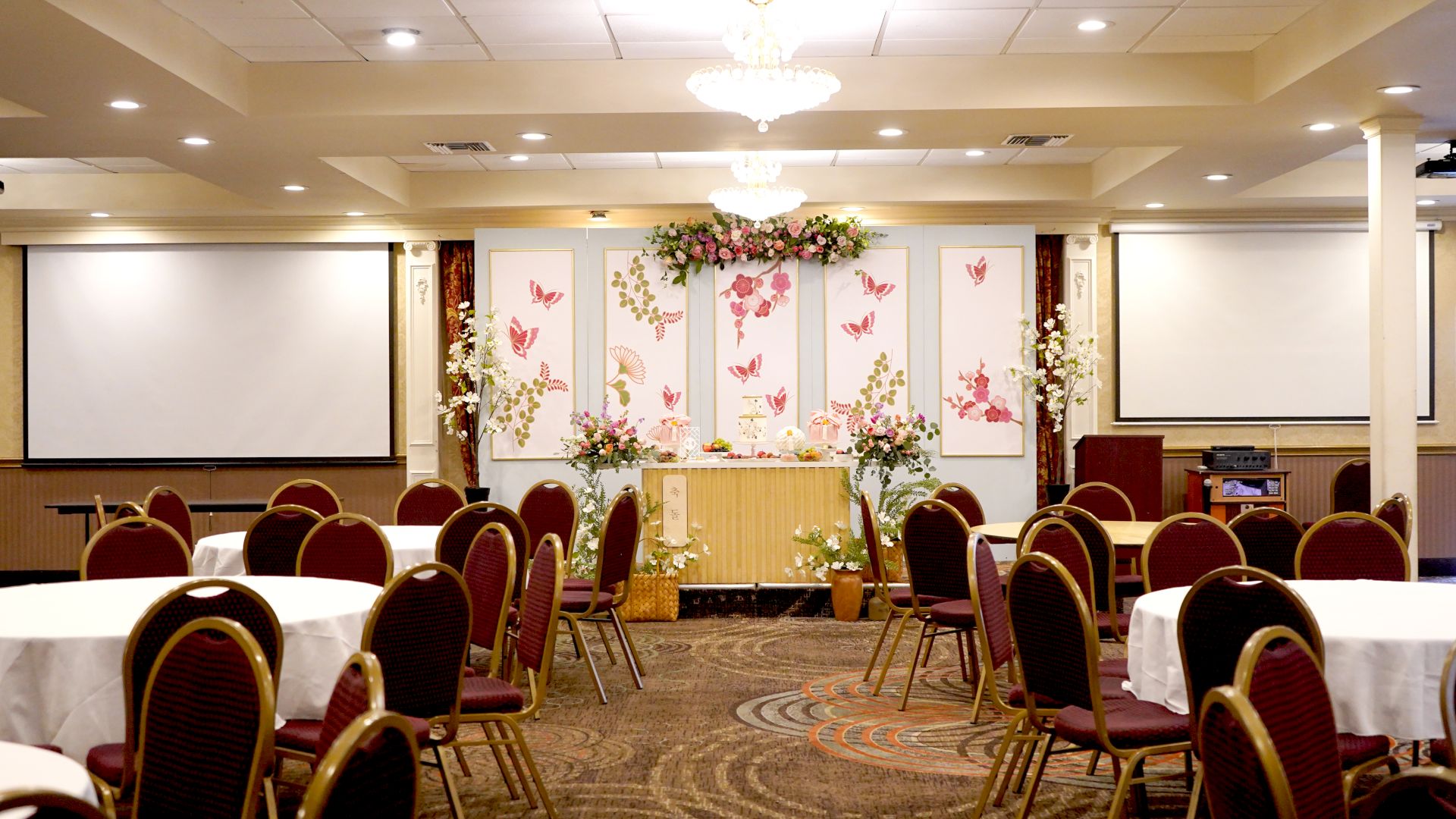 A decorated banquet hall with round tables and chairs arranged, floral arrangements, and two large projection screens on either side.