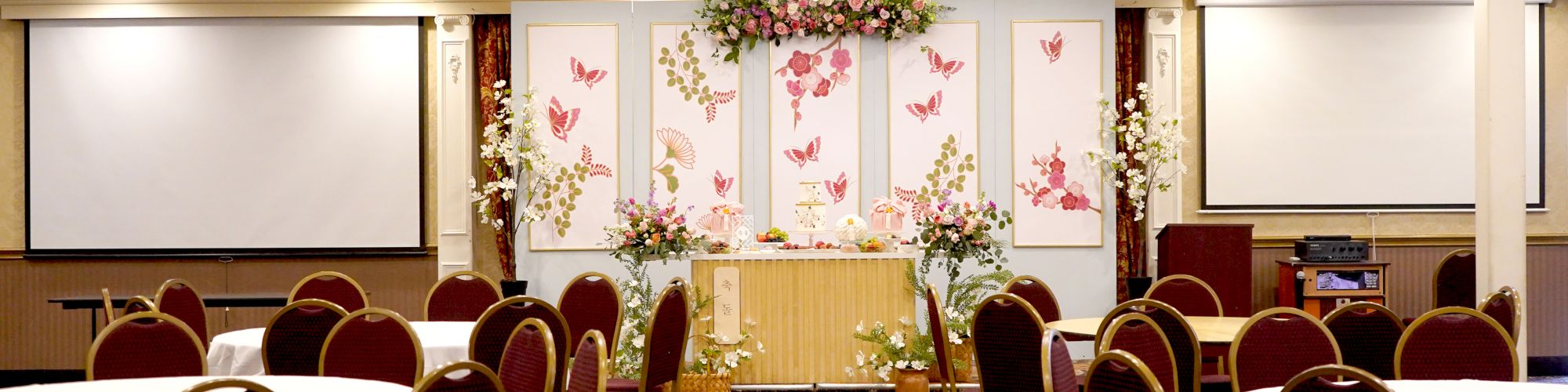 A decorated banquet hall with round tables and chairs arranged, floral arrangements, and two large projection screens on either side.