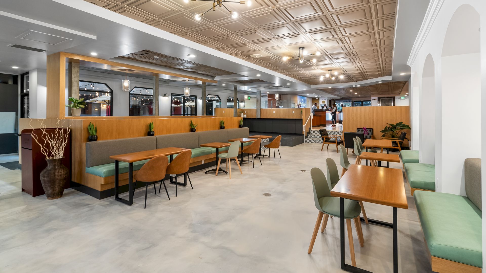 The image shows a modern, spacious dining area with wooden tables, upholstered seating, decorative lighting, and a mix of booth and chair seating.