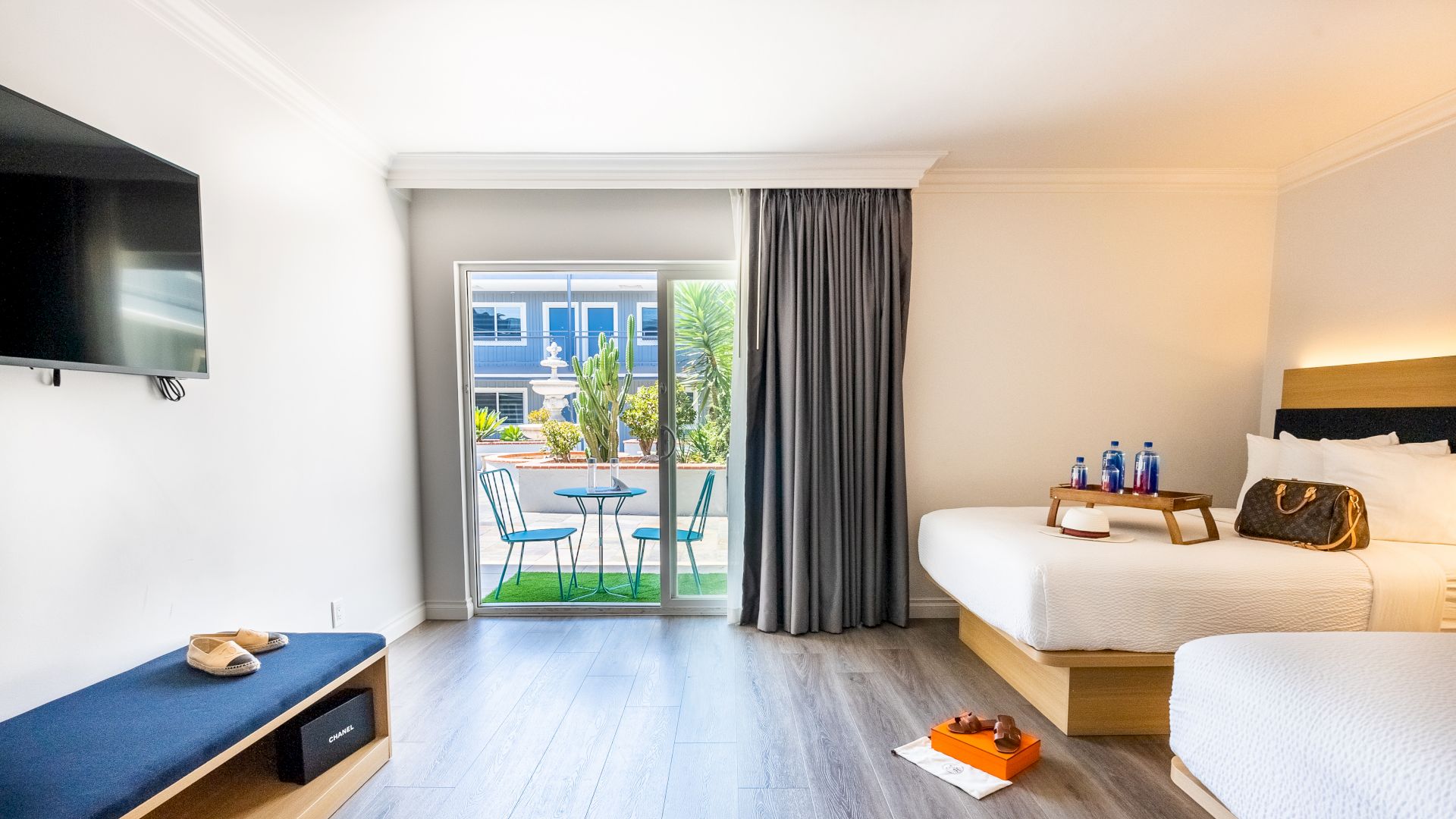A bright hotel room with two beds, TV, bench, and a sliding door leading to a patio with a table and chairs. Items on the floor and bed.