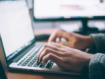 A person is typing on a laptop with another monitor in the background displaying a blurred image or website.