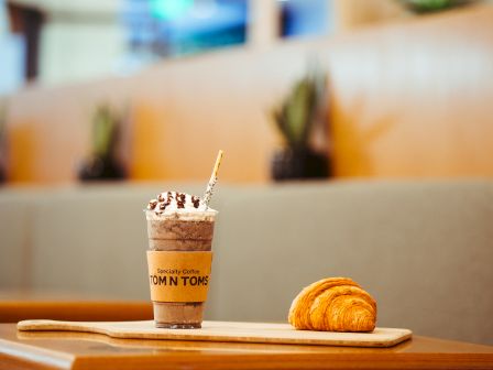 A table with a chocolate-drizzled drink and a croissant on a wooden board in a cozy café setting. Potted plants are visible in the background.