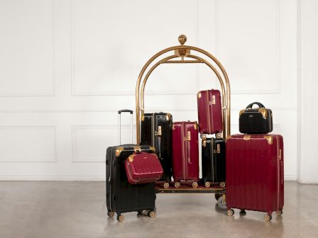 A hotel luggage cart loaded with various red and black suitcases and bags is placed in a minimally decorated room with white walls and a bare floor.