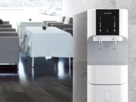 The image shows a modern white water dispenser against a marble wall, with a restaurant dining area in the background featuring tables and chairs.