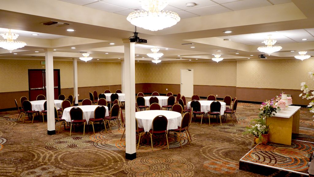 A banquet hall with round tables and chairs, elegant chandeliers, a decorated table with flowers, and carpeted flooring in a patterned design.