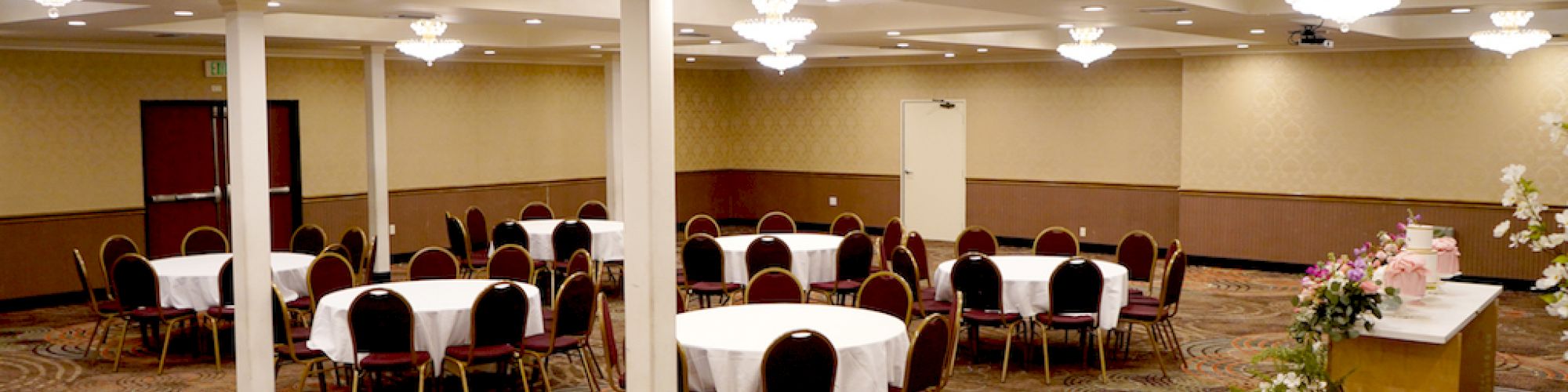 A banquet hall with round tables and chairs, elegant chandeliers, a decorated table with flowers, and carpeted flooring in a patterned design.