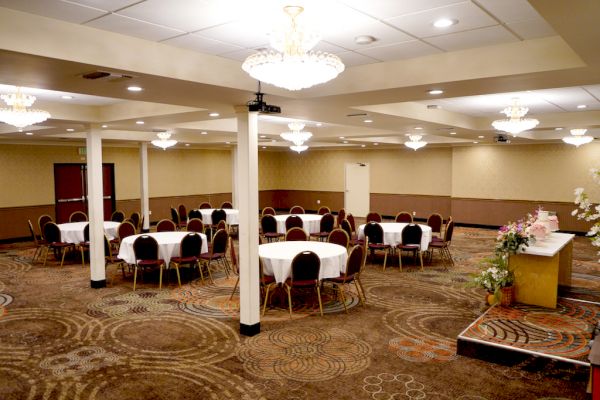 A banquet hall with round tables and chairs, chandeliers on the ceiling, decorated carpet, and a table with floral arrangements and gifts.