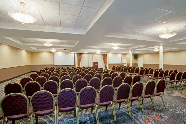 The image shows a conference room with rows of empty chairs facing a stage, a projector screen, and chandeliers hanging from the ceiling.