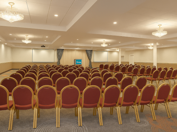 The image shows an empty conference room with rows of red chairs and a small stage at the front, under soft, ambient lighting.