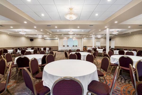 The image shows a well-lit banquet hall with several round tables covered with white cloths and surrounded by chairs, elegant chandeliers hanging.