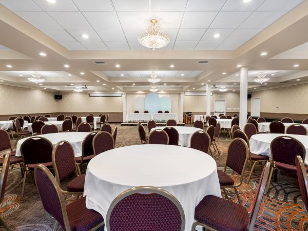 The image shows a well-lit banquet hall with several round tables covered with white cloths and surrounded by chairs, elegant chandeliers hanging.