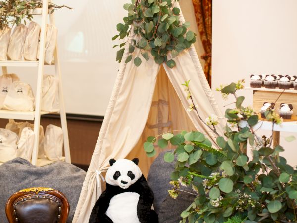 A cozy setup with a small tipi tent, a stuffed panda, a small chair, and various plants and greenery for decoration in an indoor room.