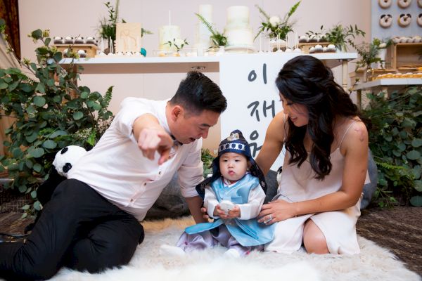 A man and woman sit on a rug with a child dressed in traditional attire. The man is pointing, and the woman is looking at the child.