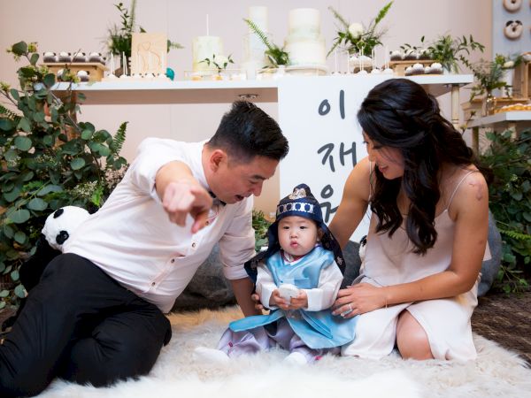 A man and woman sit on a rug with a child dressed in traditional attire. The man is pointing, and the woman is looking at the child.
