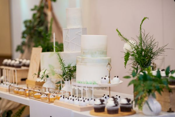 A dessert table with elegant cakes, cupcakes, and other pastries, decorated with greenery and florals, possibly for a wedding or special event.