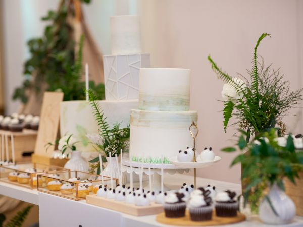 A dessert table with elegant cakes, cupcakes, and other pastries, decorated with greenery and florals, possibly for a wedding or special event.