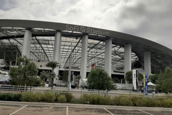 The image shows the exterior of SoFi Stadium, with visible pillars and entryways, surrounded by some greenery and an overcast sky.