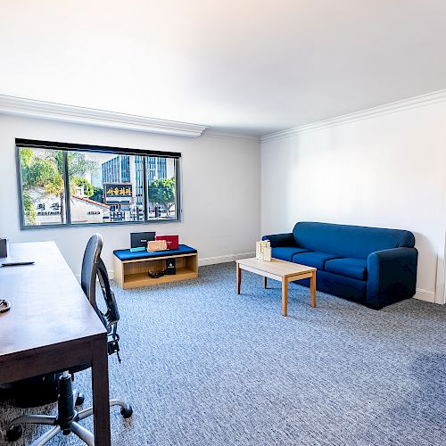 A modern, spacious living area with a desk, blue sofa, coffee table, and view of palm trees through the window, leading to a bedroom.