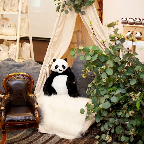 A cozy setup with a panda plush in a small tent adorned with greenery, next to a small brown chair, creating a playful and inviting atmosphere.