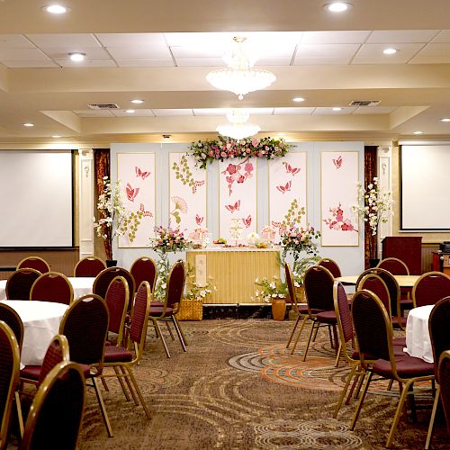 The image shows a decorated banquet hall with round tables, chairs, floral arrangements, decorative screens, and projection screens.