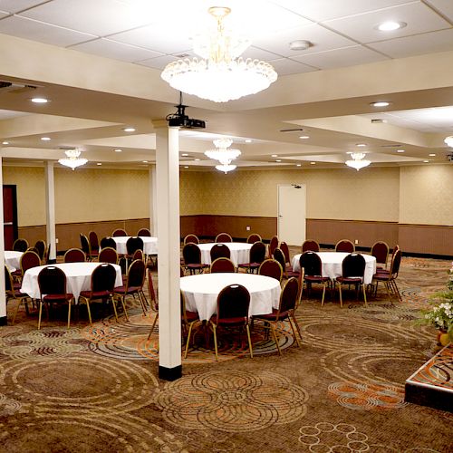 A banquet hall with round tables and chairs set up, chandeliers hanging from the ceiling, and a table with flowers on the side.